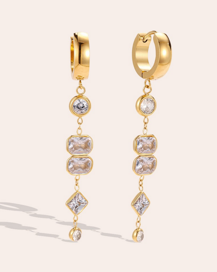Lange Ohrringe mit weißen Zirkoniasteinen von Dottilove vor beigen Hintergrund. Die Ohrringe sind gold farbig am unteren ende ist eine Kette befestigt an denen die weißen Steine hängen. 