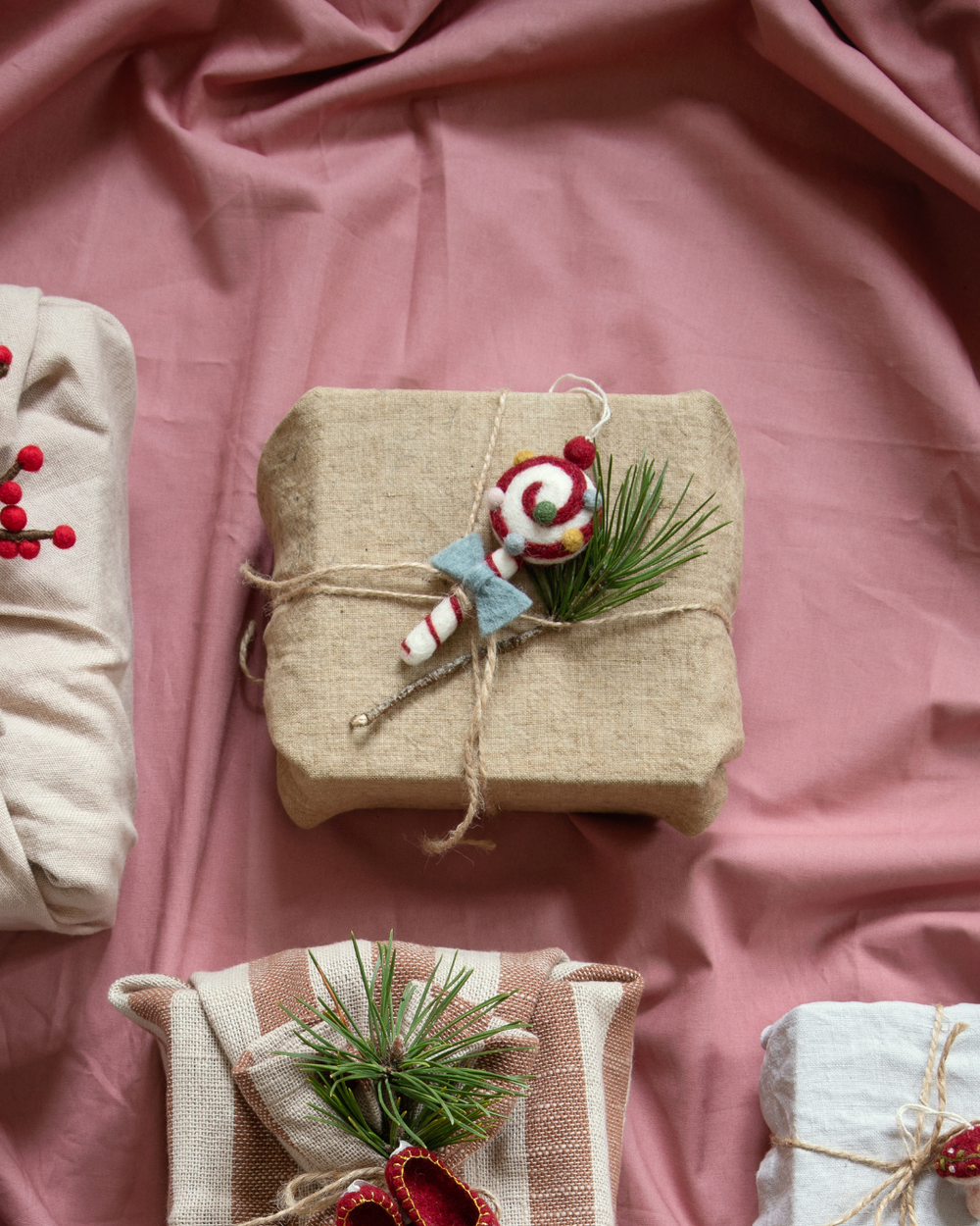 Geschenkverpackung aus Leinenstoff, dekoriert mit einem Filz-Lolli-Anhänger von Gry & Sif. Der rot-weiße Lutscher mit Spiralmuster und blauer Schleife liegt zusammen mit einem Tannenzweig auf dem naturfarbenen Paket, das mit Kordel umwickelt ist. Im Hintergrund weitere Geschenke mit weihnachtlicher Dekoration.