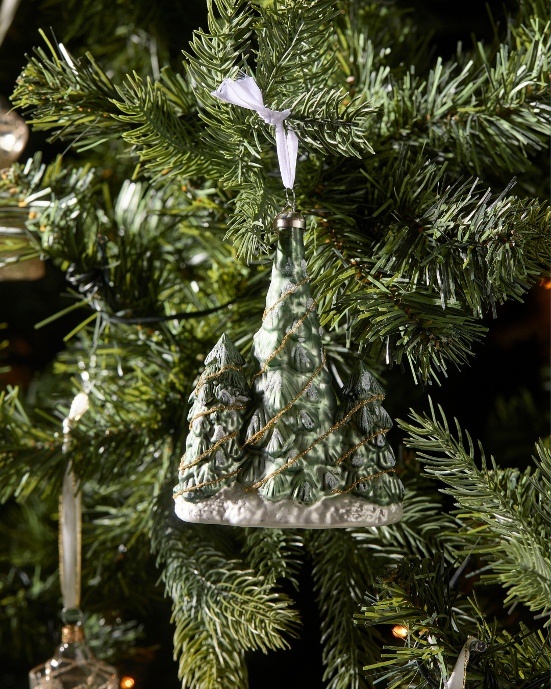 Weihnachtsbaumschmuck „Lovely Christmas Tree Ornament“ von Rivièra Maison am Tannenzweig hängend, detailreich gestaltete Tannen mit Schnee und goldener Girlande.