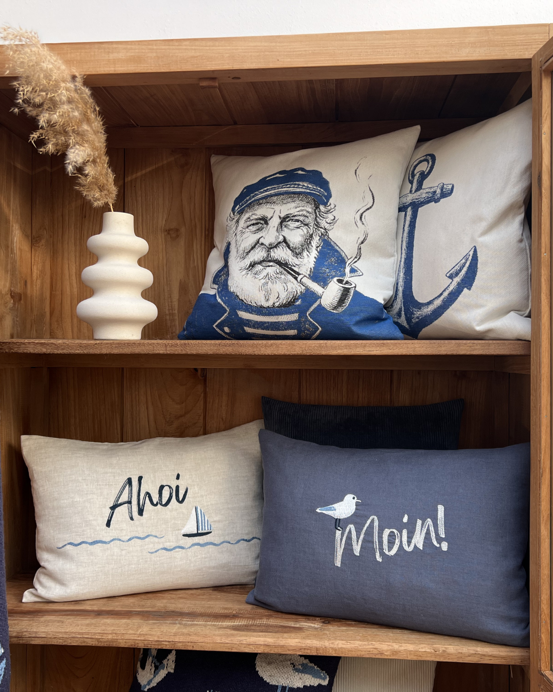 Verschiedene sommerliche Kissen von Sander sind in einem braunen Schrank auf zwei Ebenen dekoriert. Oben sind die Kissenbezüge Old Salt und Hook mit einem Seemann- und Anker-Motiv zu sehen. Darunter sind die Leinen-Kissen Ahoi und Moin dekoriert.