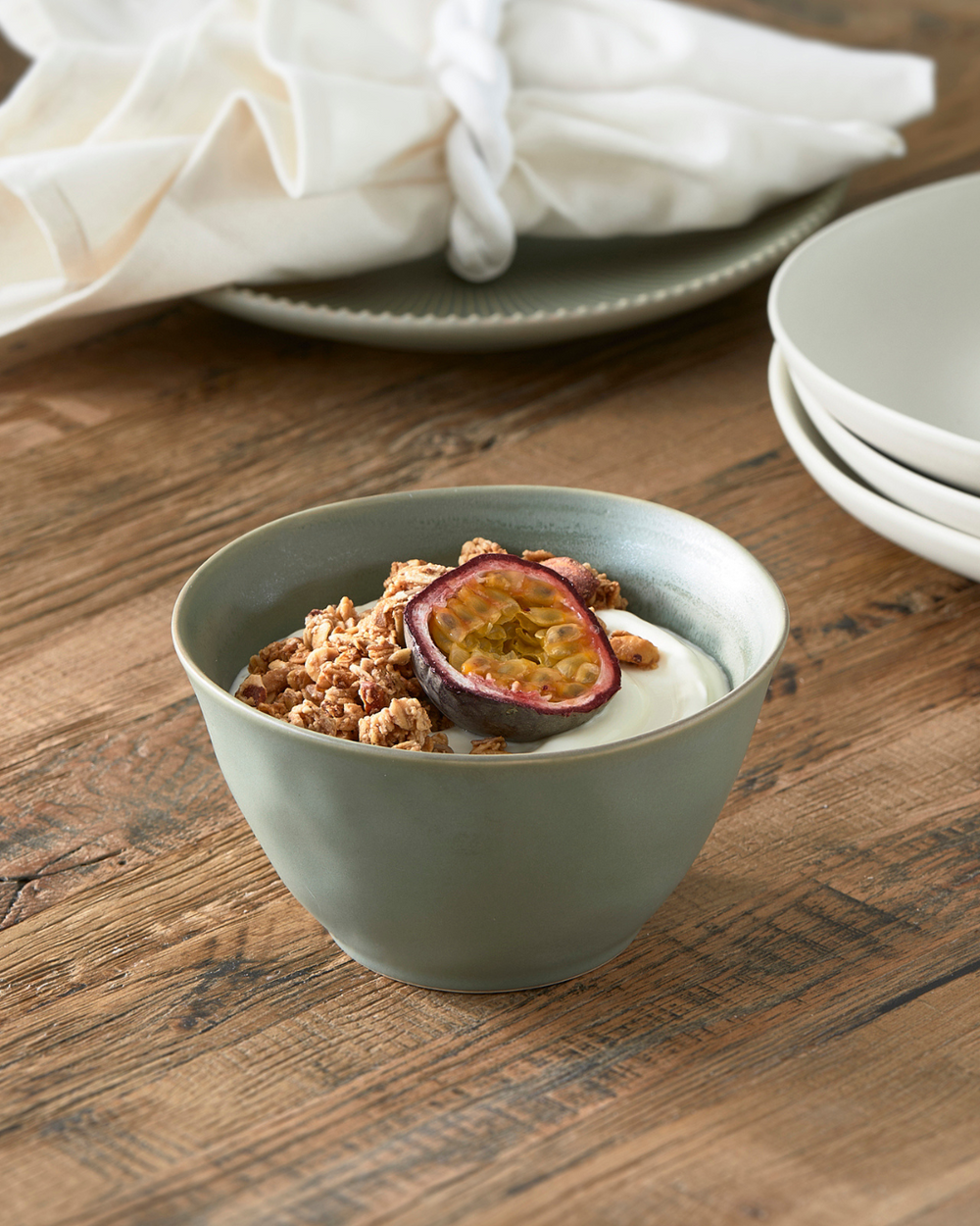 Grüne Marseille Schüssel von Rivièra Maison, dekoriert mit Joghurt, Granola und Passionsfrucht, auf rustikalem Holztisch mit passendem Geschirr im Hintergrund.