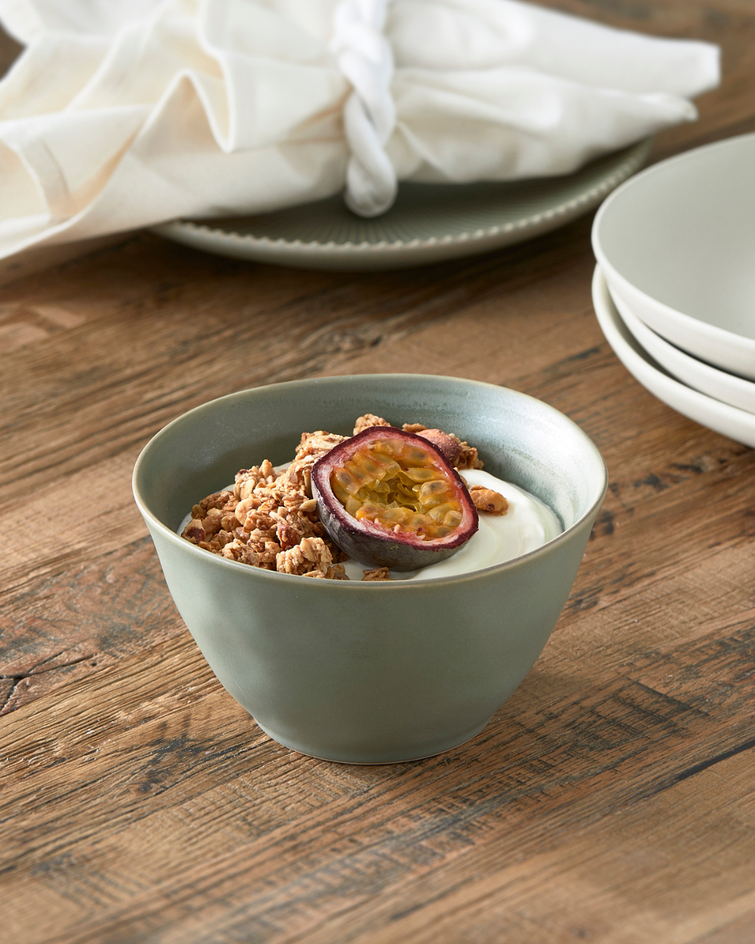 Grüne Marseille Schüssel von Rivièra Maison, dekoriert mit Joghurt, Granola und Passionsfrucht, auf rustikalem Holztisch mit passendem Geschirr im Hintergrund.