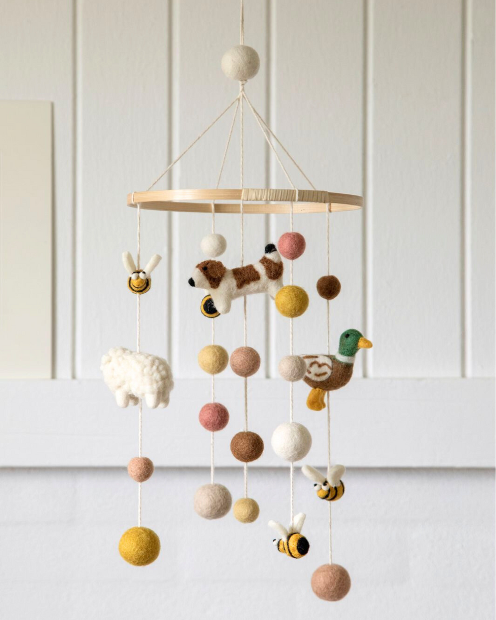 Das Bauernhof Mobile von Gry & Sif mit Hund, Ente, Schaf, Bienen und bunten Filzkugeln vor beigefarbener Wand im Kinderzimmer.