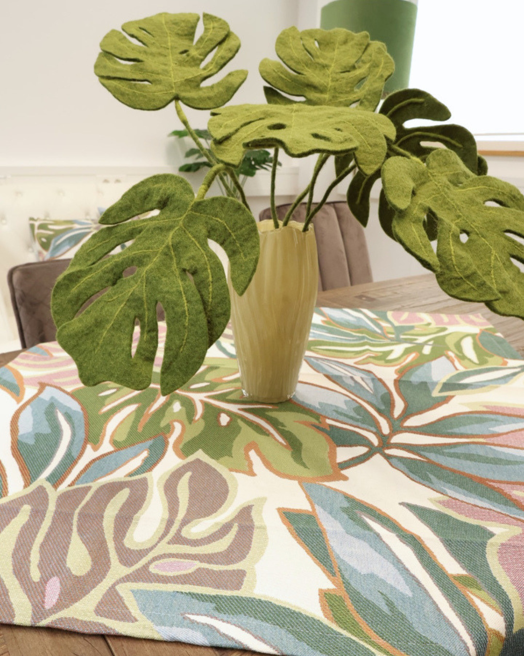 Zimmerpflanzen Mitteldecke Selva von Sander Table + Home. Die mehrfarbigen Monsterblätter sind sich gegenseitig überlappend dargestellt. Blaue, grüne und roséfarbene Blätter mit sich farblich absetzender Blattumrandung sind in den Stoff der Mitteldecke gewoben. Auf der Decke steht eine Vase, welche mit Monsterablättern aus grünem Filz gefüllt ist.