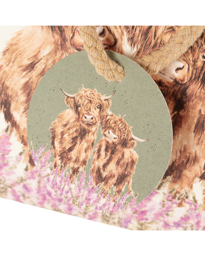 Nahansicht auf die Geschenktasche und den dazugehörigen Patch. Die Geschenktasche zeigt zwei flauschige Hochlandrinder mit brauner Fellfarbe, die in einem Feld aus violett-rosanen Fingerhutblumen stehen. Im für Hannah Dale typischen natürlichen Stil blicken die beiden sanftmütig über die Blüten hinweg. Die Tasche ist am oberen Rand mit einer Schleife verschlossen. Ein Patch, welcher an der Verschlusskordel angebracht ist zeigt ebenfalls das Hochlandkuh-Motiv.