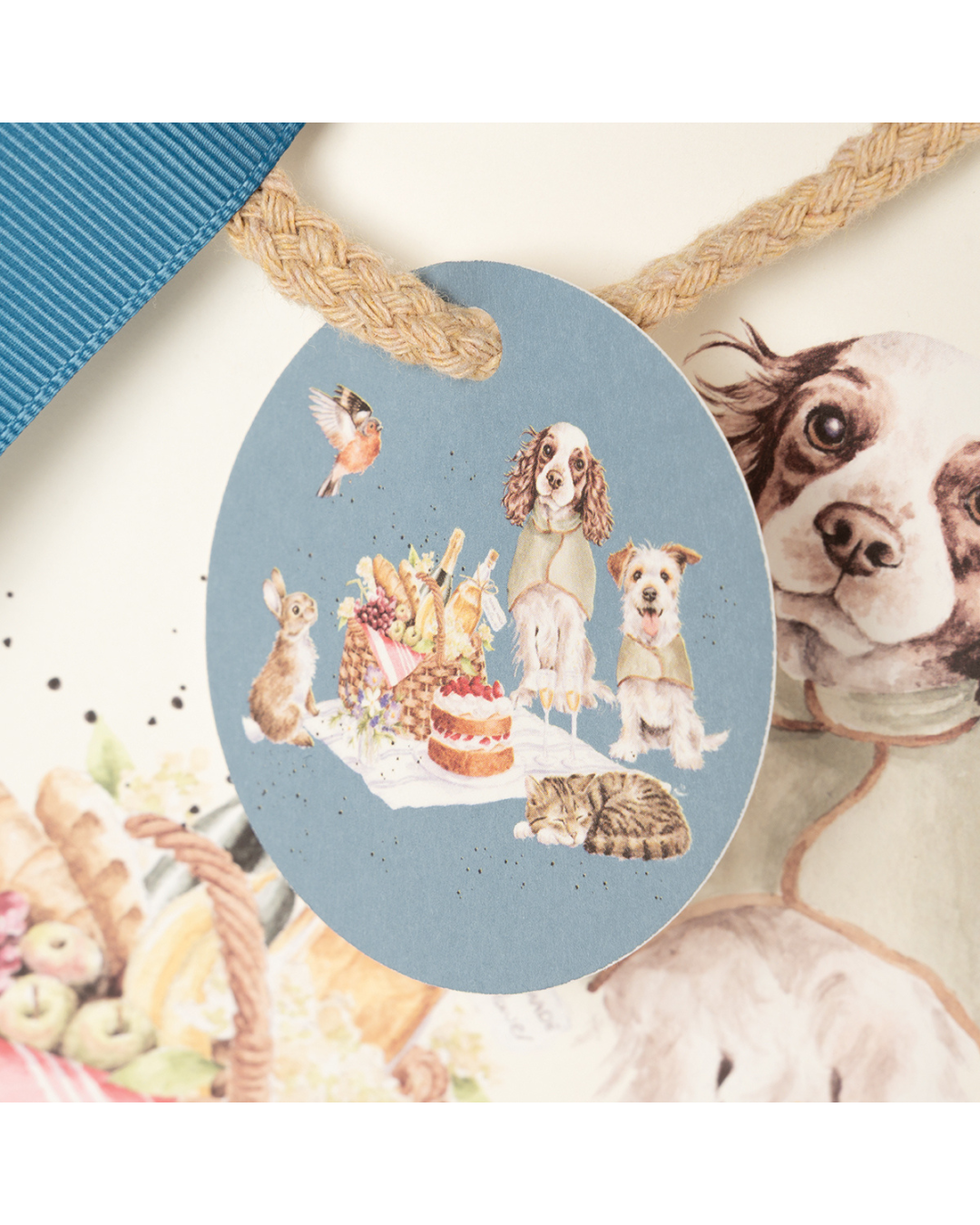 Nahansicht Geschenktasche "The Hoppy Picnic" von Wrendale Designs. Zu sehen ist eine Illustration von zwei Hunden mit Überwurfdeckchen und ein Kaninchen sitzend auf einer Picknickdecke. Vor der Picknickdecke liegt eine braun-schwarz getigerte schlafende Katze. Ein niedliches Rotkehlchen fliegt über die Picknickdecke hinweg, auf welcher sich ein prall gefüllter Picknickkorb und ein Erbeerkuchen befinden. Die Darstellung befindet sich auf einem an einer Kordel befestigten Schildchen an der Geschenktasche.
