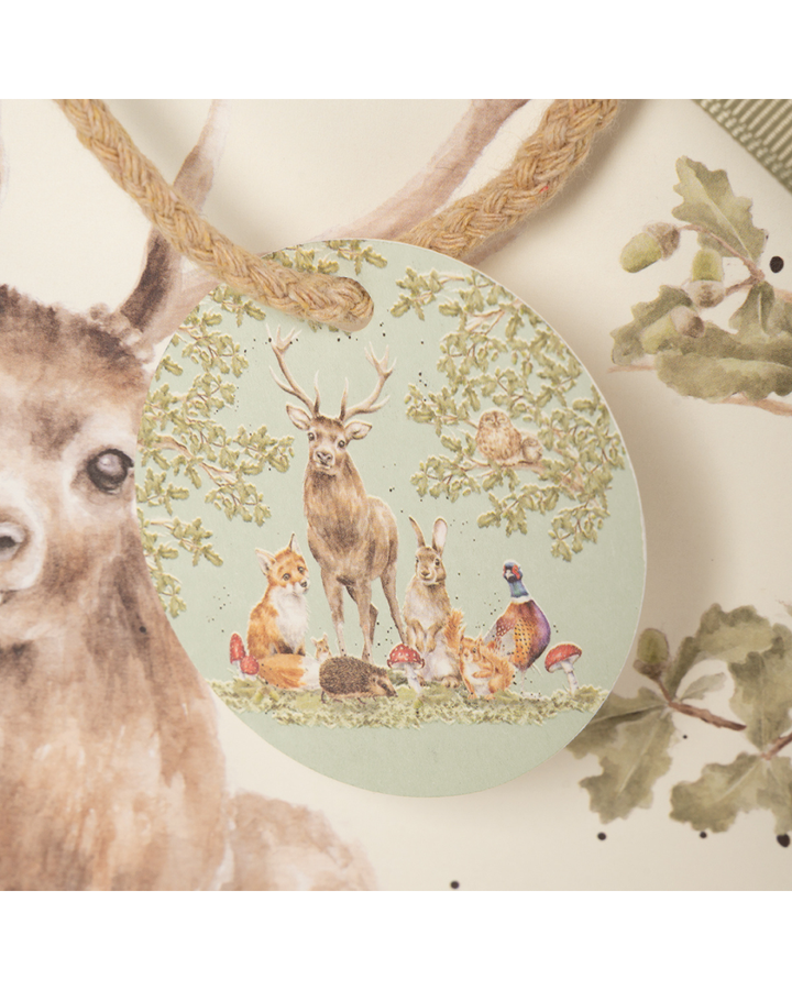 Die Geschenktasche zeigt die Bewohner des Waldes im typisch-natürlichen Stil von Hannah Dale. Hirsch, Fuchs, Hase, Igel, Eichhörnchen, Maus und Fasan stehen unter Eichenbäumen und Zweigen. In der Eiche selbst sitzt ein niedlicher Kauz mit noch süßerem Nachwuchs. Das detailreiche Motiv wird durch Insekten, wie Hummel und Schmetterling, sowie Eicheln und Fliegenpilze vervollständigt. Hier zu sehen ist der Patch, welcher an der Tütenverschlusskordel befestigt ist. dieser zeigt ebenfalls das Waldtier-Motiv.