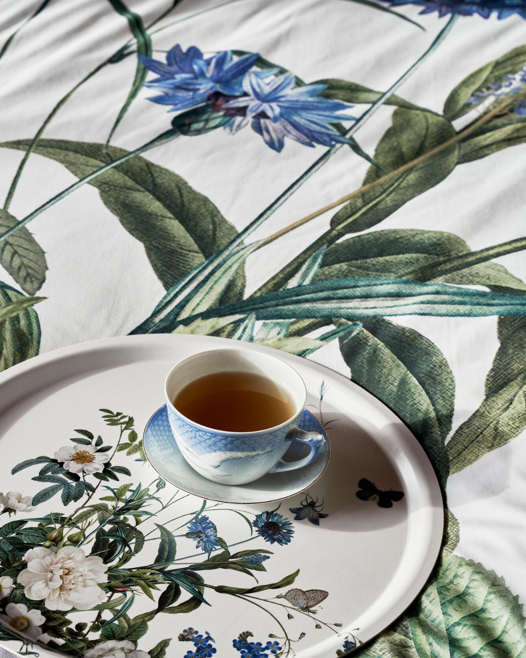 Tasse Tee auf einem Tablett mit Blumenmotiven, platziert auf der Bettwäsche „Blue Flower Garden“ von Koustrup & Co., Nahaufnahme der blauen und weißen Blumen.