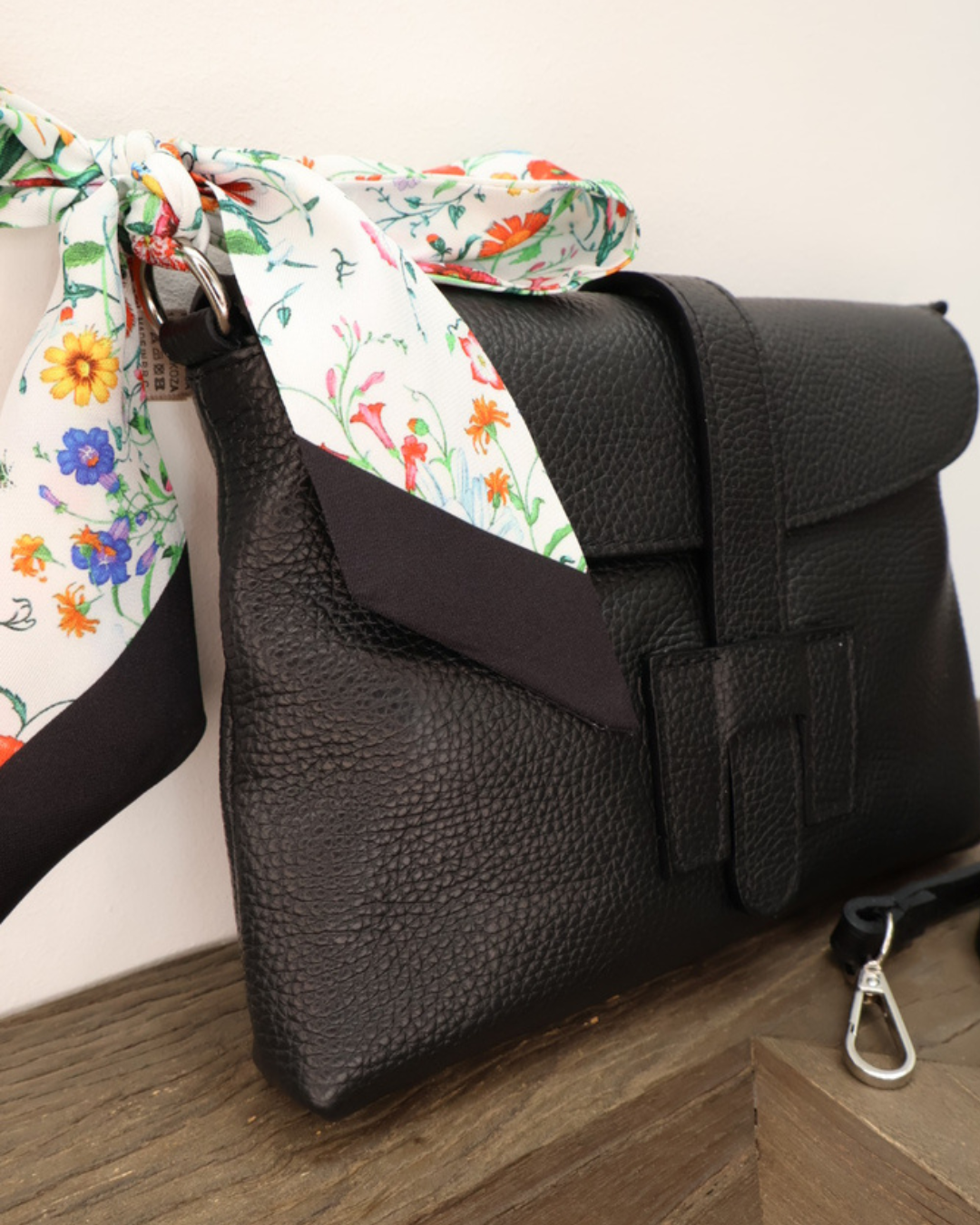 Nahansicht der Ledertasche "Isabella" vom Südstrand in schwarz. Die Tasche liegt auf einem Holztisch vor einem weißen Hintergrund während das bunte florale-Bandana gut zu sehen ist.