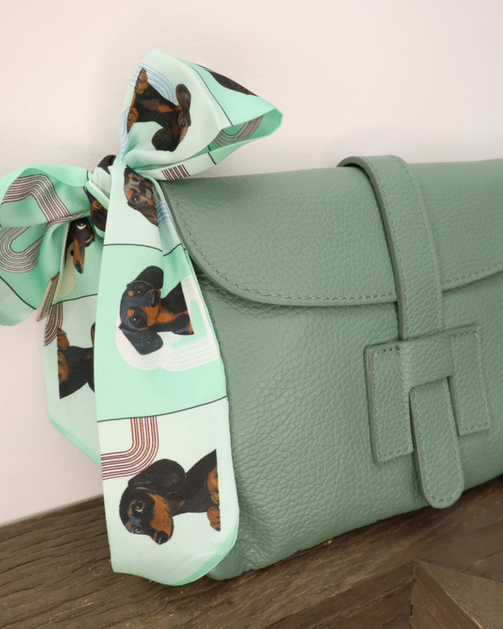 Nahansicht der Ledertasche "Isabella" vom Südstrand in Seegras grün. Die Tasche liegt auf einem Holztisch vor einem weißen Hintergrund während das grüne Dackel-Bandana gut zu sehen ist.