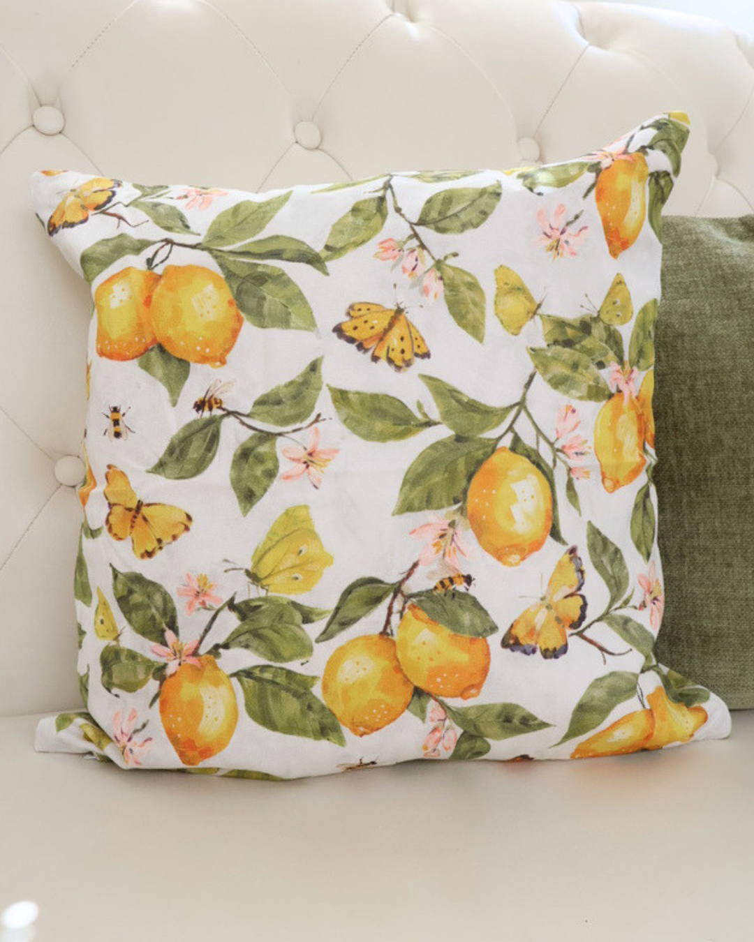 Nahaufnahme des Zitronenkissens "Fresh Lemons"  von Ambiente. Das Motiv ist größtenteils gelb und grün. Zu sehen sind Schmetterlinge, Zitronen, Blumen und Bienen. Das Kissen liegt auf einem weißen Sofa.