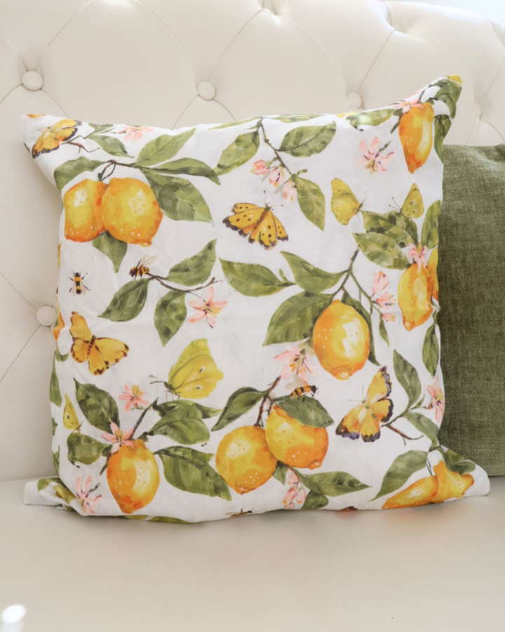 Nahaufnahme des Zitronenkissens "Fresh Lemons"  von Ambiente. Das Motiv ist größtenteils gelb und grün. Zu sehen sind Schmetterlinge, Zitronen, Blumen und Bienen. Das Kissen liegt auf einem weißen Sofa.