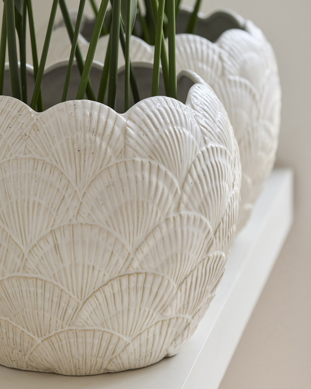 Blumentopf mit Muschelmuster von Rivièra Maison, gefüllt mit Palmlilie, auf weißem Tisch.