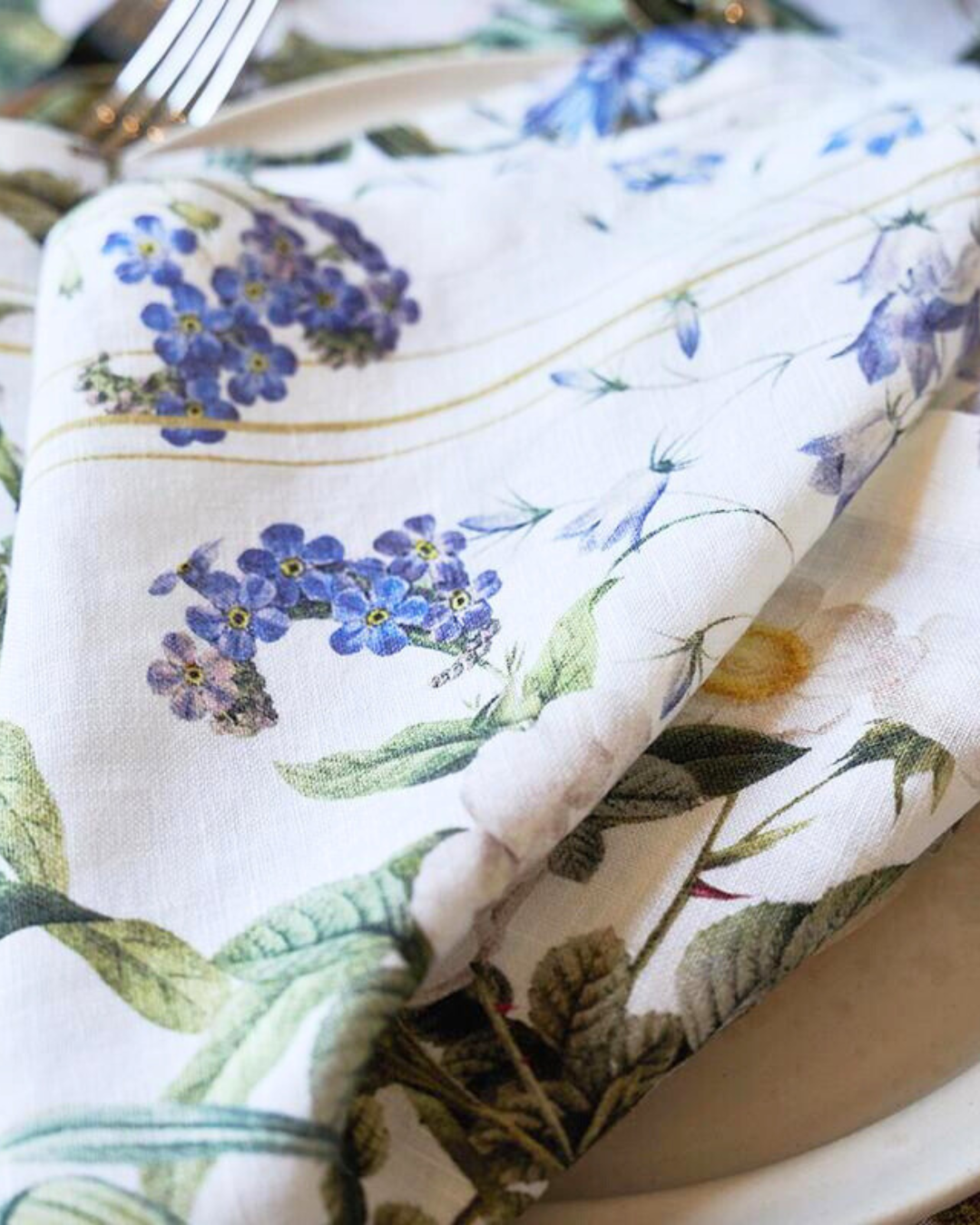 Eine Leinenserviette „Blue Flower Garden“ von Koustrup & Co., leicht gefaltet, liegt auf einem weißen Teller, umgeben von Besteck und dekorativen Elementen.