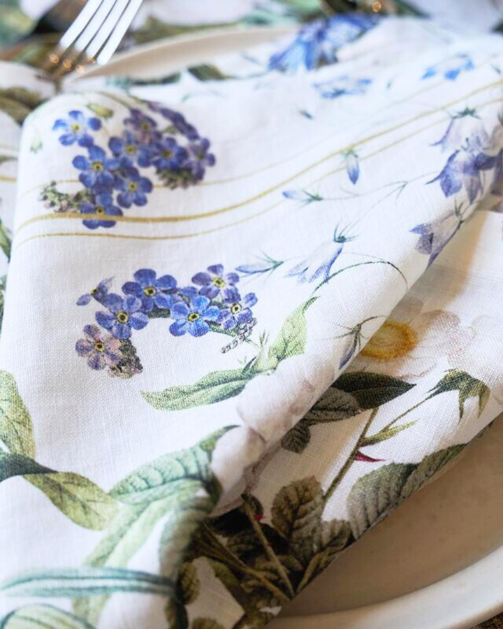 Eine Leinenserviette „Blue Flower Garden“ von Koustrup & Co., leicht gefaltet, liegt auf einem weißen Teller, umgeben von Besteck und dekorativen Elementen.