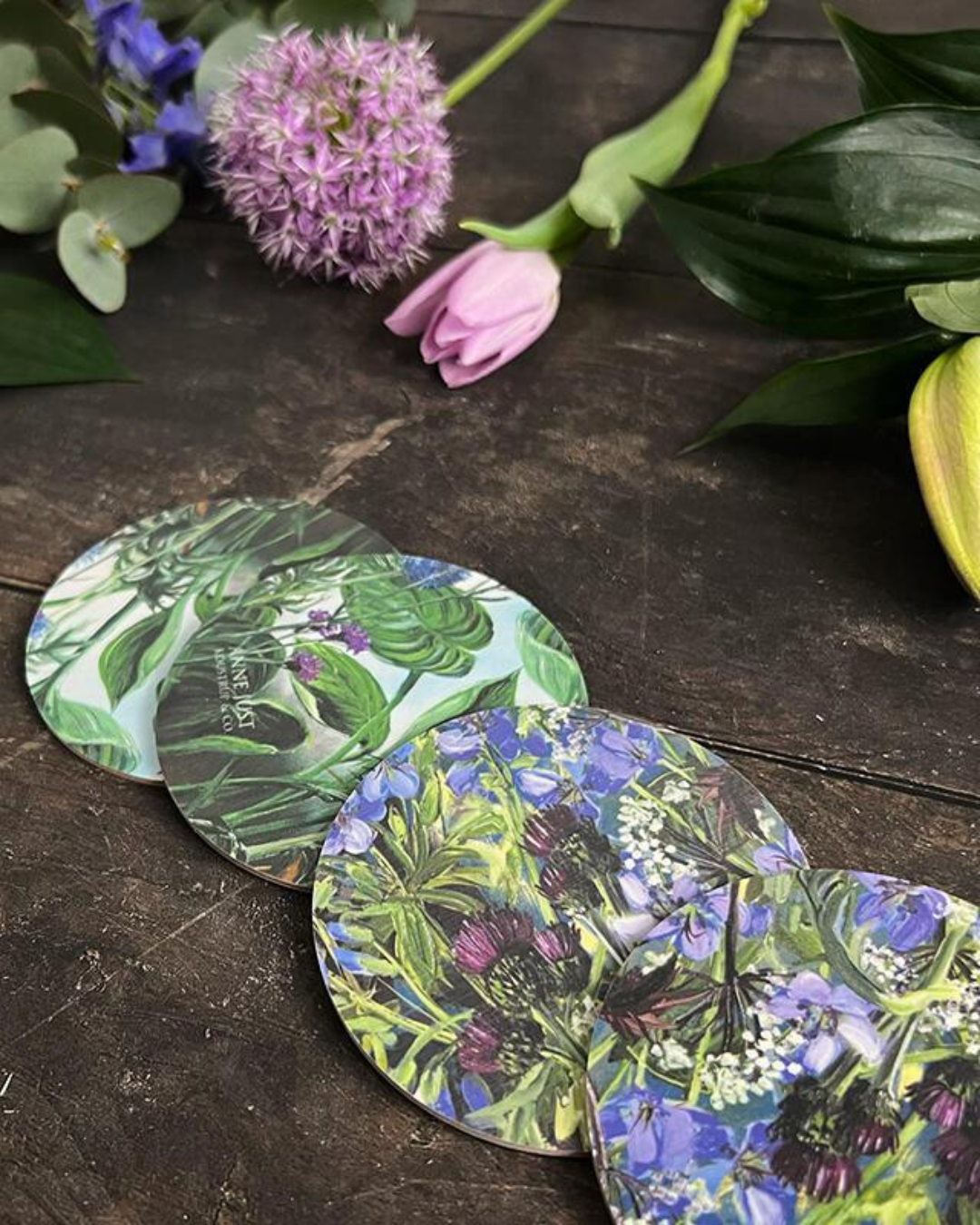 4er Set "Anne Just" Untersetzer von Koustrup & co auf dunklen Holztisch umgeben von echten Blumen.