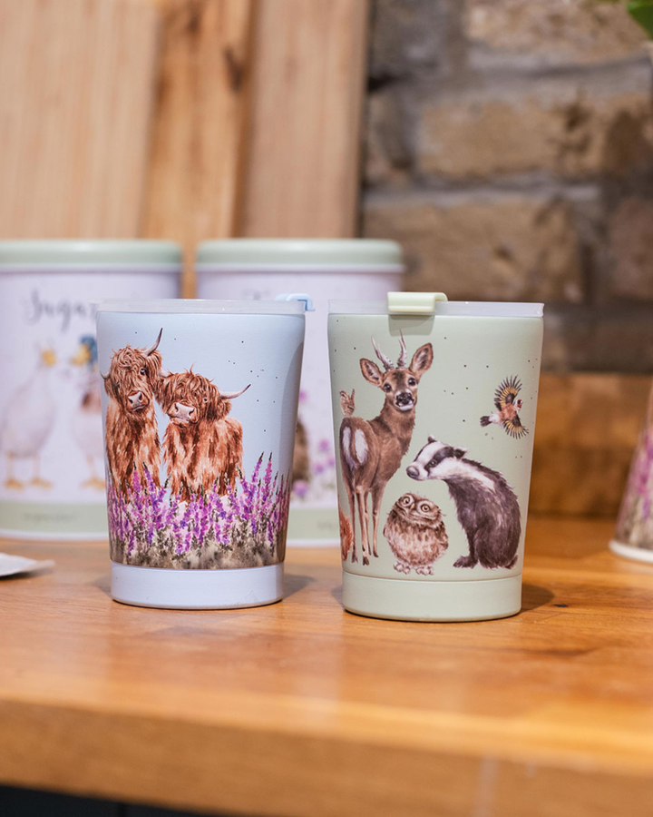 Thermobecher To Go von Wrendale Designs. Auf der linken Seite steht der Thermobecher "Highland Cow" und rechts daneben der Thermobecher "Woodland". Die natürlichen Motive von Hannah Dale zeigen zum Einen zwei Hochlandrinder auf einer Blühwiese und zum Anderen einige der verschiedenen Waldbewohner.