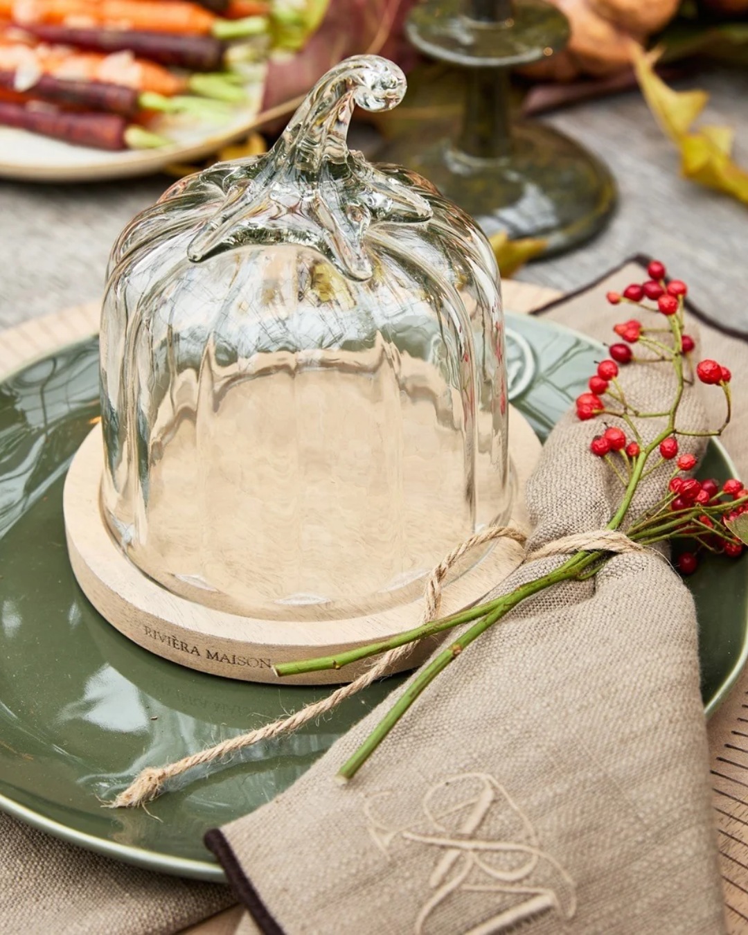 Kürbis Glasglocke "Pumpkin Cloche" von Riviera Maison mit Holzuntersetzer mit schwarzer RM Aufschrift. Hier in Großaufnahme auf einem gedeckten Tisch, auf einem Teller stehend.