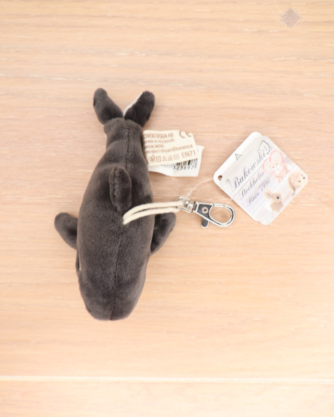 Der Orca Schlüsselanhänger Oreo von Bukowski liegt auf einem Holztisch. Der Metallkarabiner und das Markenetikett „Bukowski Stockholm Since 1990“ sind sichtbar.