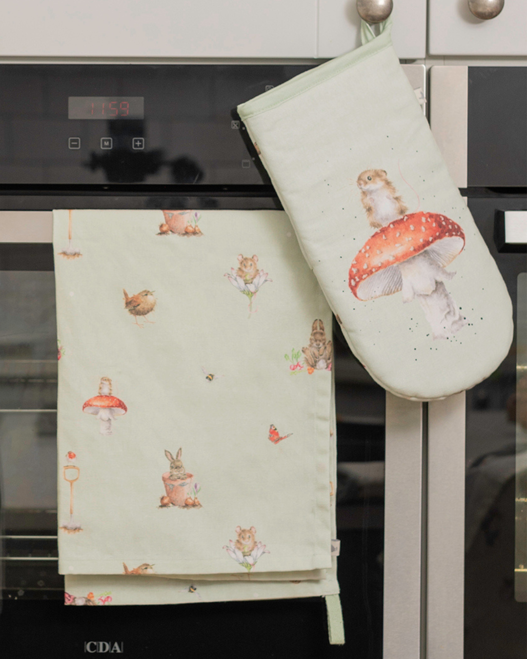 Ein Ofenhandschuh mit der Maus auf dem Fliegenpilz und ein passendes Geschirrtuch mit den Gartentieren von Wrendale Designs drauf, hängen an einem Backofen.
