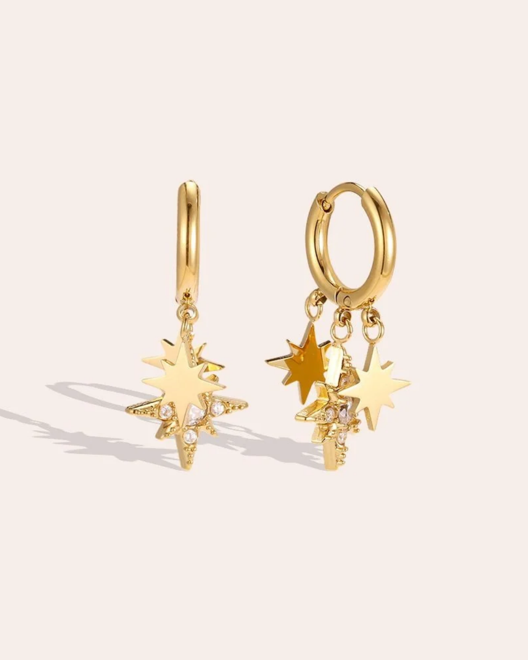 Ohrringe mit Sternen von Dottilove vor beigen Hintergrund. Die Ohrringe sind golden farbig mit 3 kleinen Sternanhängern. Einer ist groß mit weißen Steinen.