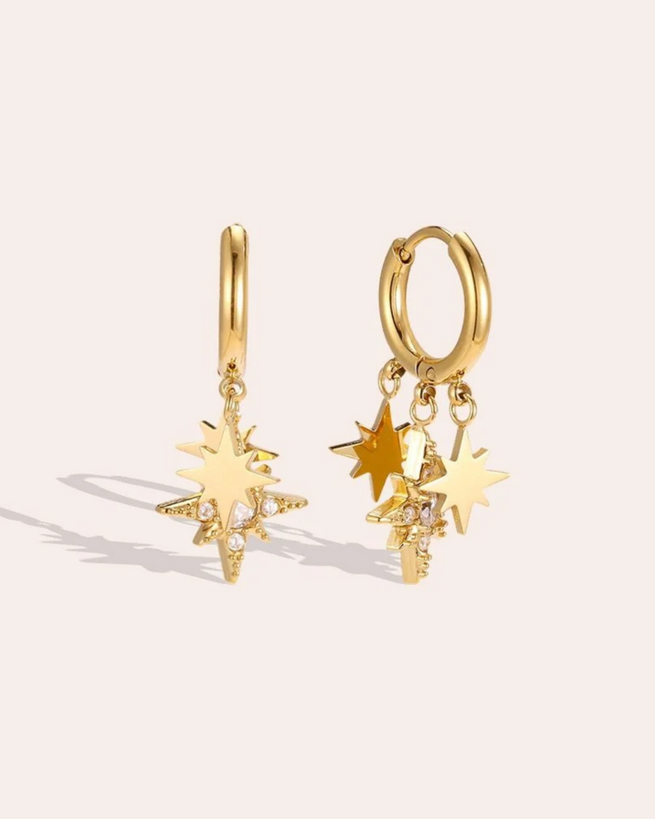 Ohrringe mit Sternen von Dottilove vor beigen Hintergrund. Die Ohrringe sind golden farbig mit 3 kleinen Sternanhängern. Einer ist groß mit weißen Steinen.