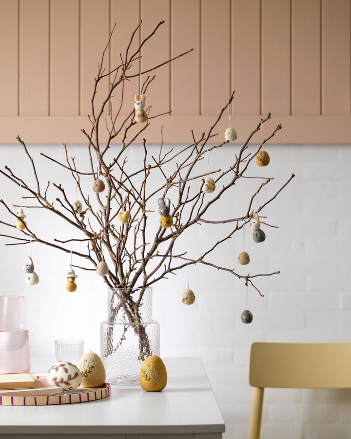 Großer Osterstrauch in einer Glasvase, dekoriert mit verschiedenen Filz-Ostereiern und Hasen-Anhängern auf einem Tisch.