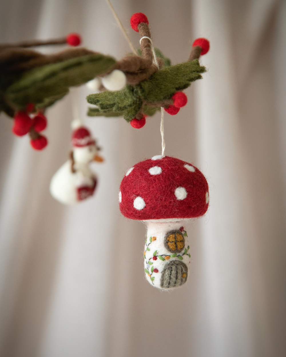 Weihnachtsanhänger Pilzhaus aus Filz von Gry & Sif, handgefertigt in Nepal, zeigt ein märchenhaftes Häuschen in Form eines Fliegenpilzes mit rotem Dach und weißen Punkten, einem kleinen runden Fenster und einer Tür, dekoriert mit grünen Ranken und roten Beeren.