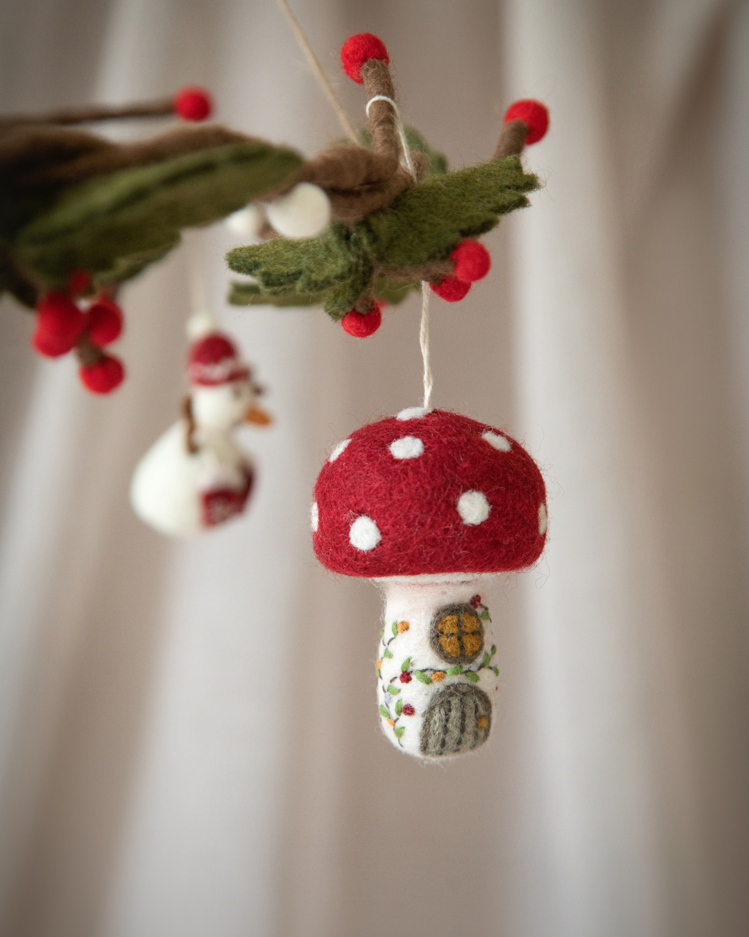 Weihnachtsanhänger Pilzhaus aus Filz von Gry & Sif, handgefertigt in Nepal, zeigt ein märchenhaftes Häuschen in Form eines Fliegenpilzes mit rotem Dach und weißen Punkten, einem kleinen runden Fenster und einer Tür, dekoriert mit grünen Ranken und roten Beeren.