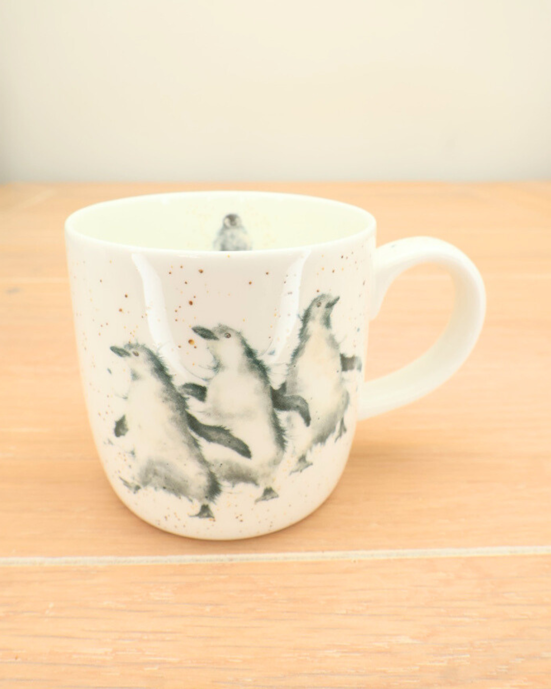 Die Pinguin Tasse "Out on the Town" von Wrendale Designs steht auf einem Holztisch. Das Motiv zeigt drei Pinguine auf hellem Grund.