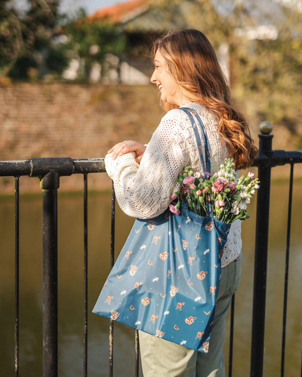 Dunkelblaue Kuh Einkaufstasche Awakening von Wrendale Designs im Gebrauch. Eine junge Dame steht an einem Geländer und blickt lächelnd auf einen Fluss. Um ihre Schulter trägt sie die Einkaufstasche, aus welcher die Köpfe von bunten Blumen ragen.