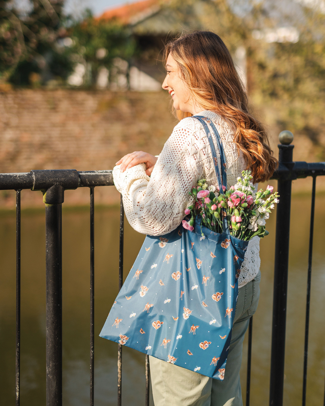 Dunkelblaue Kuh Einkaufstasche Awakening von Wrendale Designs im Gebrauch. Eine junge Dame steht an einem Geländer und blickt lächelnd auf einen Fluss. Um ihre Schulter trägt sie die Einkaufstasche, aus welcher die Köpfe von bunten Blumen ragen.