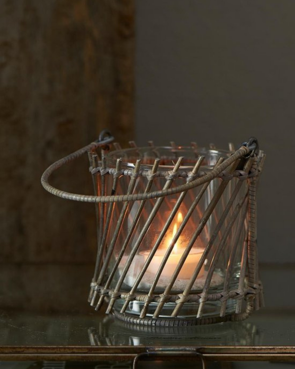 Stmmungsvoll leuchtendes Dinard Windlicht aus Rattan von Rivièra Maison auf einem Glastisch.