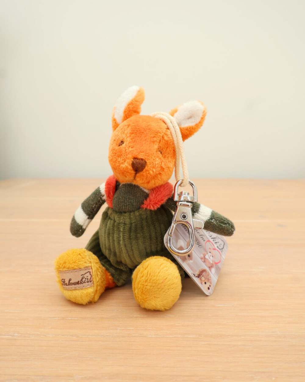 Ein orangefarbenes Stofftier-Kaninchen mit grünem Pullover sitzt auf einem hellen Holztisch. Es trägt einen silbernen Karabiner mit Etikett an der Hand und hat gelbe Pfoten mit dem Markenaufdruck „Bukowski“.