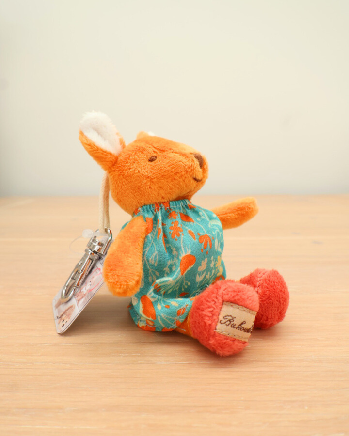 Das orangefarbene Kaninchen-Stofftier trägt ein türkisfarbenes Kleid mit Blumenmuster und sitzt auf einem Holztisch. Es hält einen silbernen Karabiner mit Etikett in der Hand, und seine Pfoten sind knallrot.