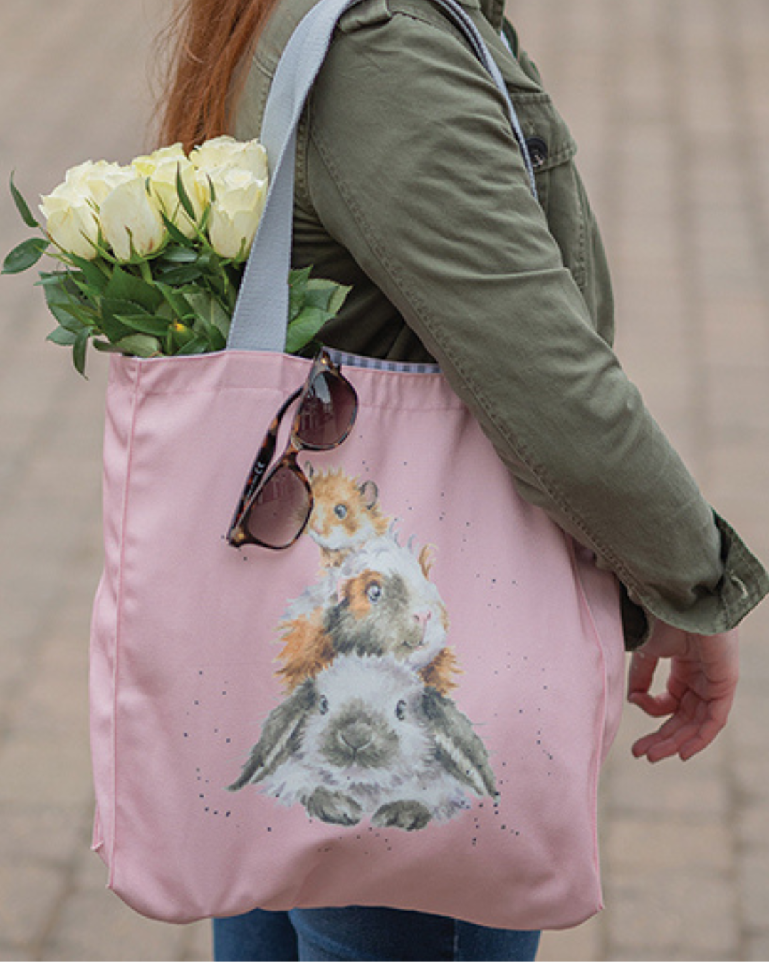 Rosa Nager Einkaufstasche von Wrendale Designs getragen von einer Frau. Zu sehen ist ein Hamster, ein Meerschweinchen und ein Kaninchen auf einander liegend der Größe nach sortiert. Aus der Tasche hängen weiße Tulpen und eine Sonnenbrille.
