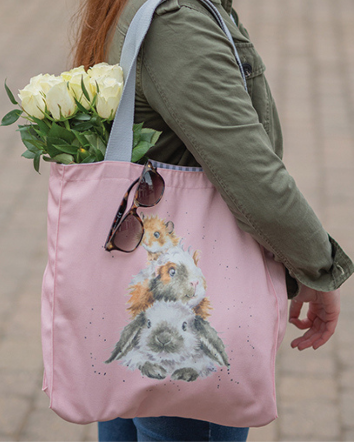 Rosa Nager Einkaufstasche von Wrendale Designs getragen von einer Frau. Zu sehen ist ein Hamster, ein Meerschweinchen und ein Kaninchen auf einander liegend der Größe nach sortiert. Aus der Tasche hängen weiße Tulpen und eine Sonnenbrille.