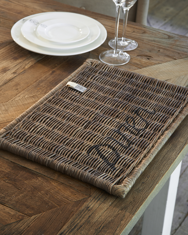 Das Rustic Rattan Diner Platzset von Rivièra Maison liegt auf einem Esstisch mit brauner Holzplatte und weißen Tischbeinen.