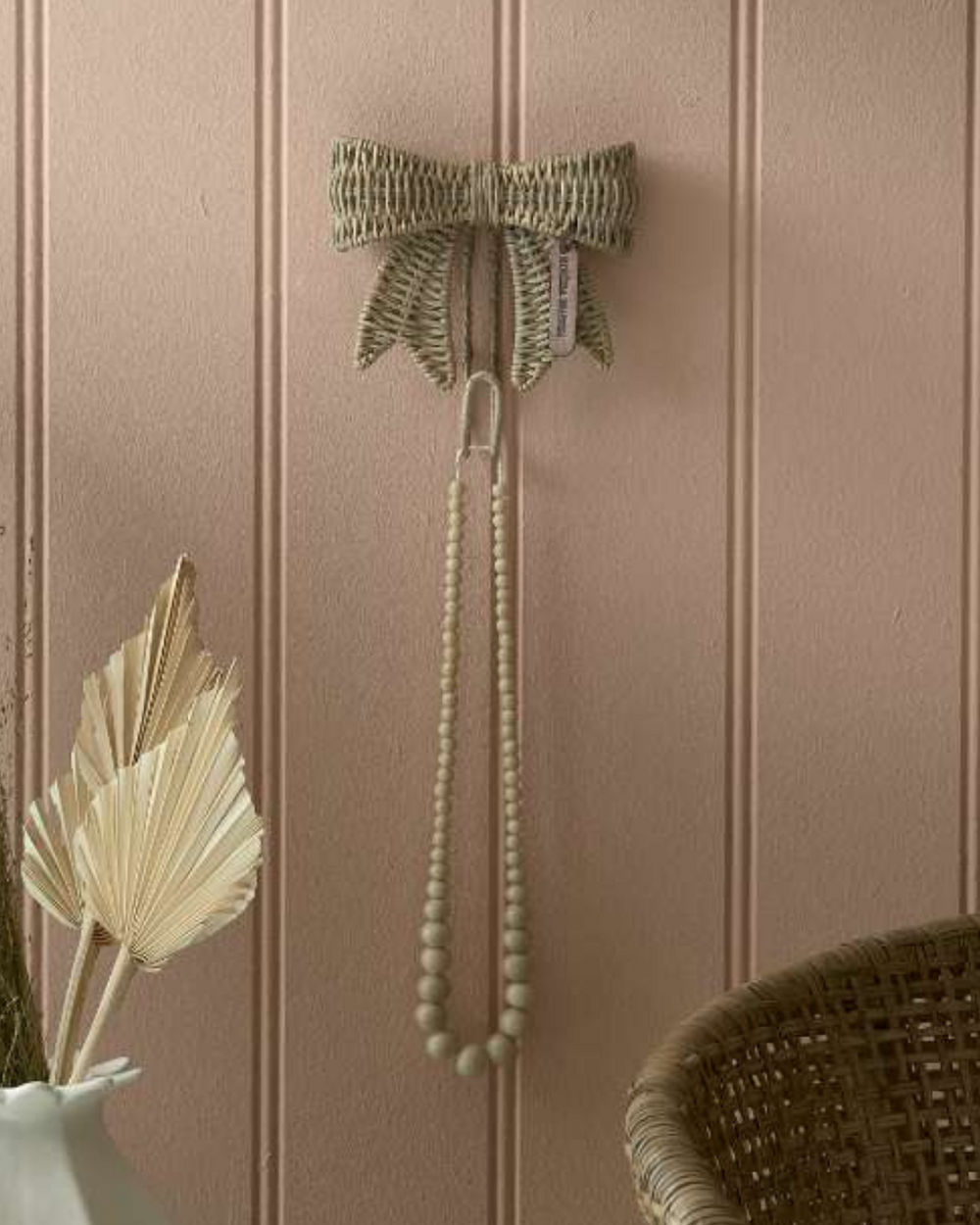 Rustic Rattan Schleifenhaken von Rivièra Maison an rosafarbener Wand mit Holzperlenkette als Dekoration.