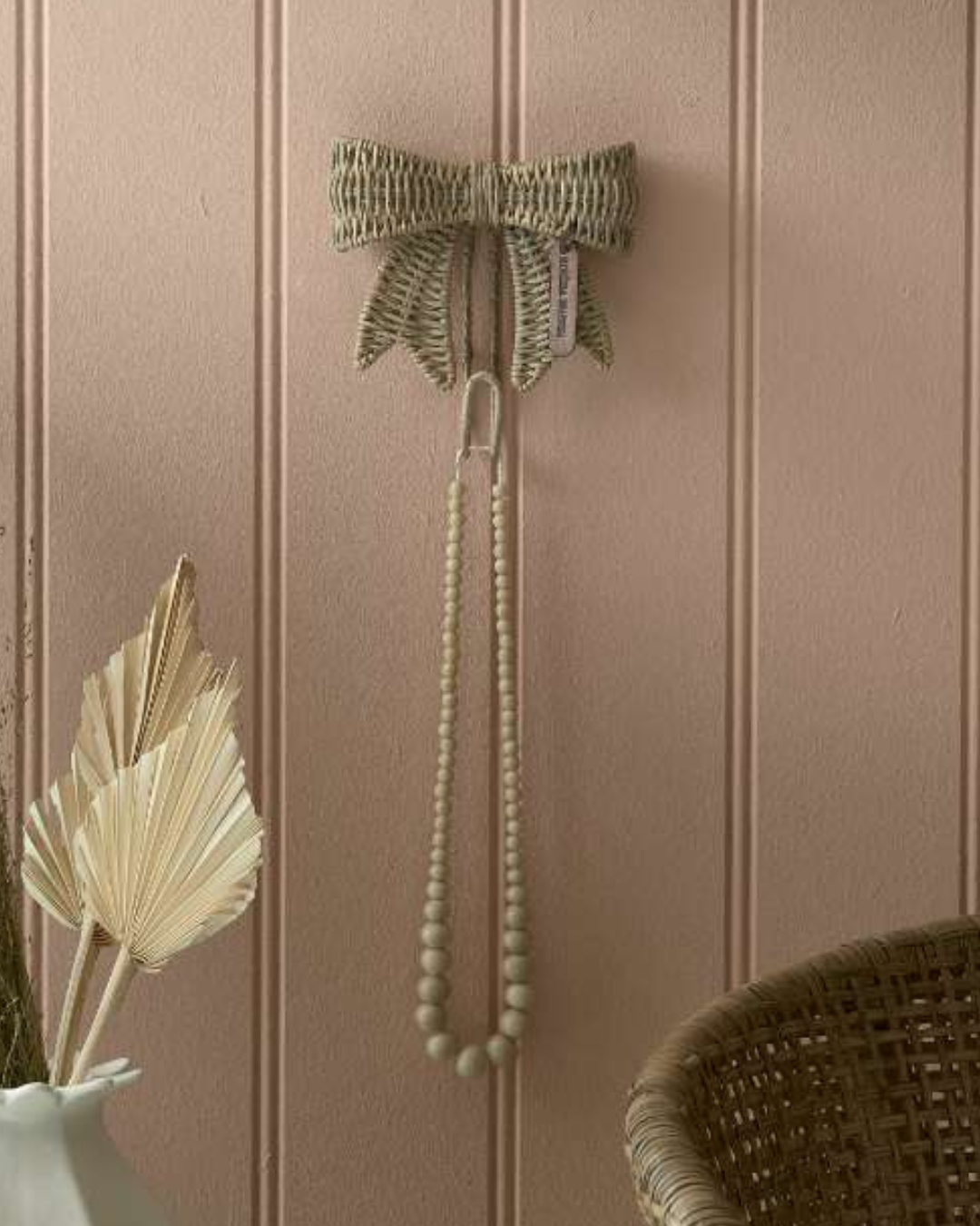 Rustic Rattan Schleifenhaken von Rivièra Maison an rosafarbener Wand mit Holzperlenkette als Dekoration.