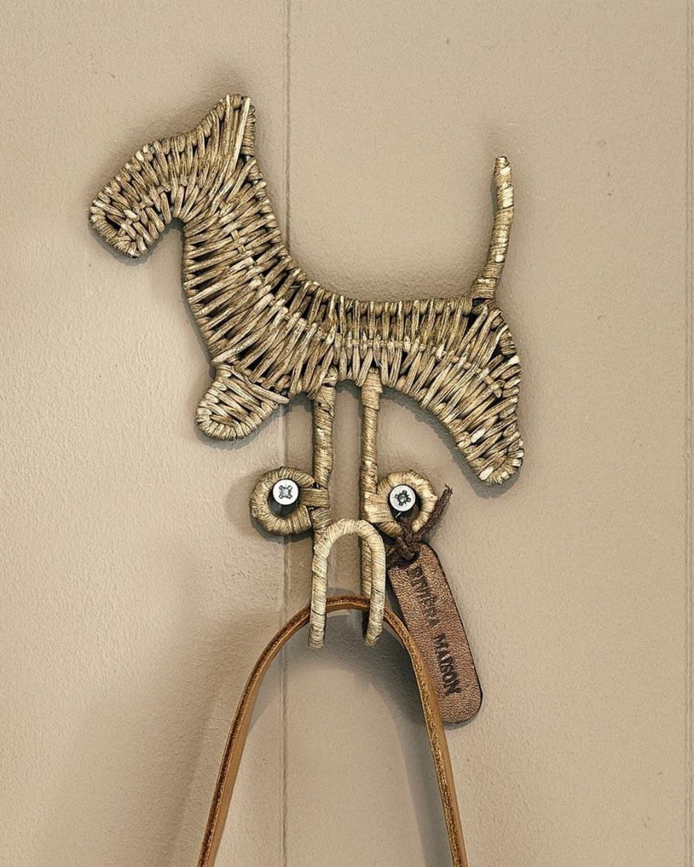 Rustic Rattan Hunde Haken von Rivièra Maison vor beiger Wand mit Hundeleine dran.