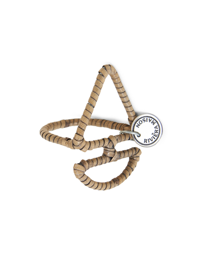 Ein handgefertigter Serviettenring aus braunem Rattan in Form eines Segelschiffs. An der Seite ist ein rundes Metall-Emblem mit der Aufschrift „Rivièra Maison“ befestigt. Der Ring liegt freigestellt auf weißem Hintergrund und zeigt die filigrane Verarbeitung.