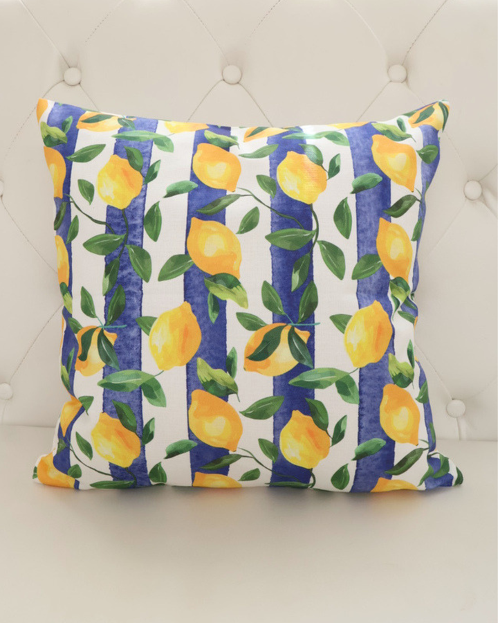Bild des Zitronen Kissens "Lemontree". Das Motiv besteht aus Blättern und Zitronen vor einem Blau Weiß gestreiften Hintergrund. Das Kissen liegt auf einem weißen Sofa.