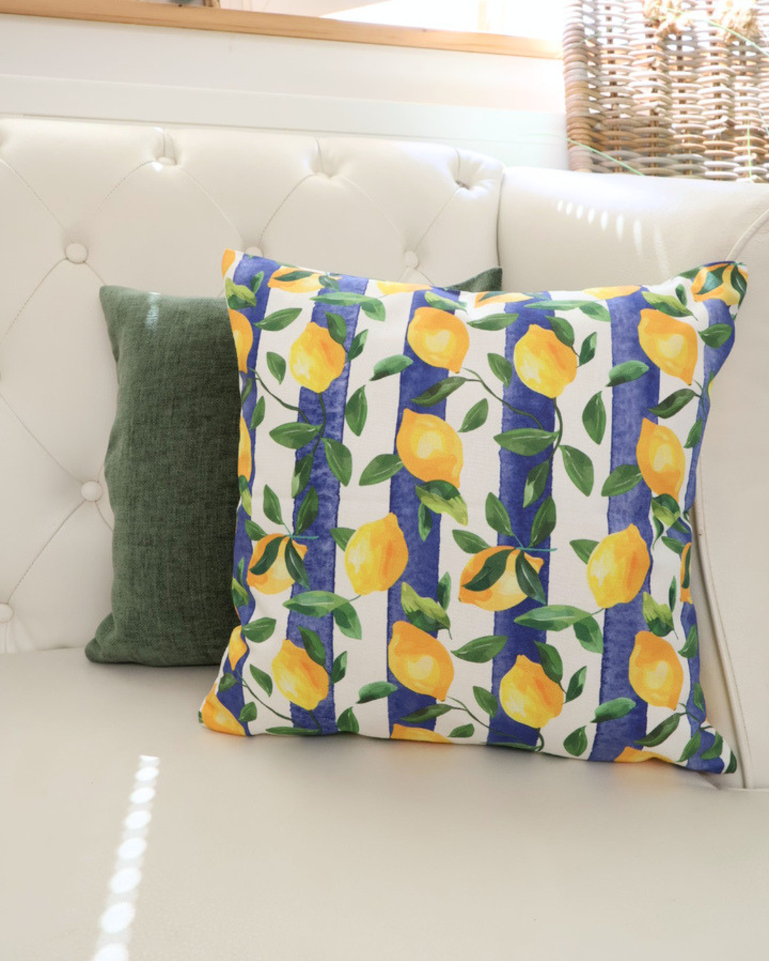 Dekoiertes Bild des Zitronen Kissens "Lemontree". Das Motiv besteht aus Blättern und Zitronen vor einem Blau Weiß gestreiften Hintergrund. Das Kissen liegt neben dem "Kensignton" Kissen auf einem weißen Sofa.