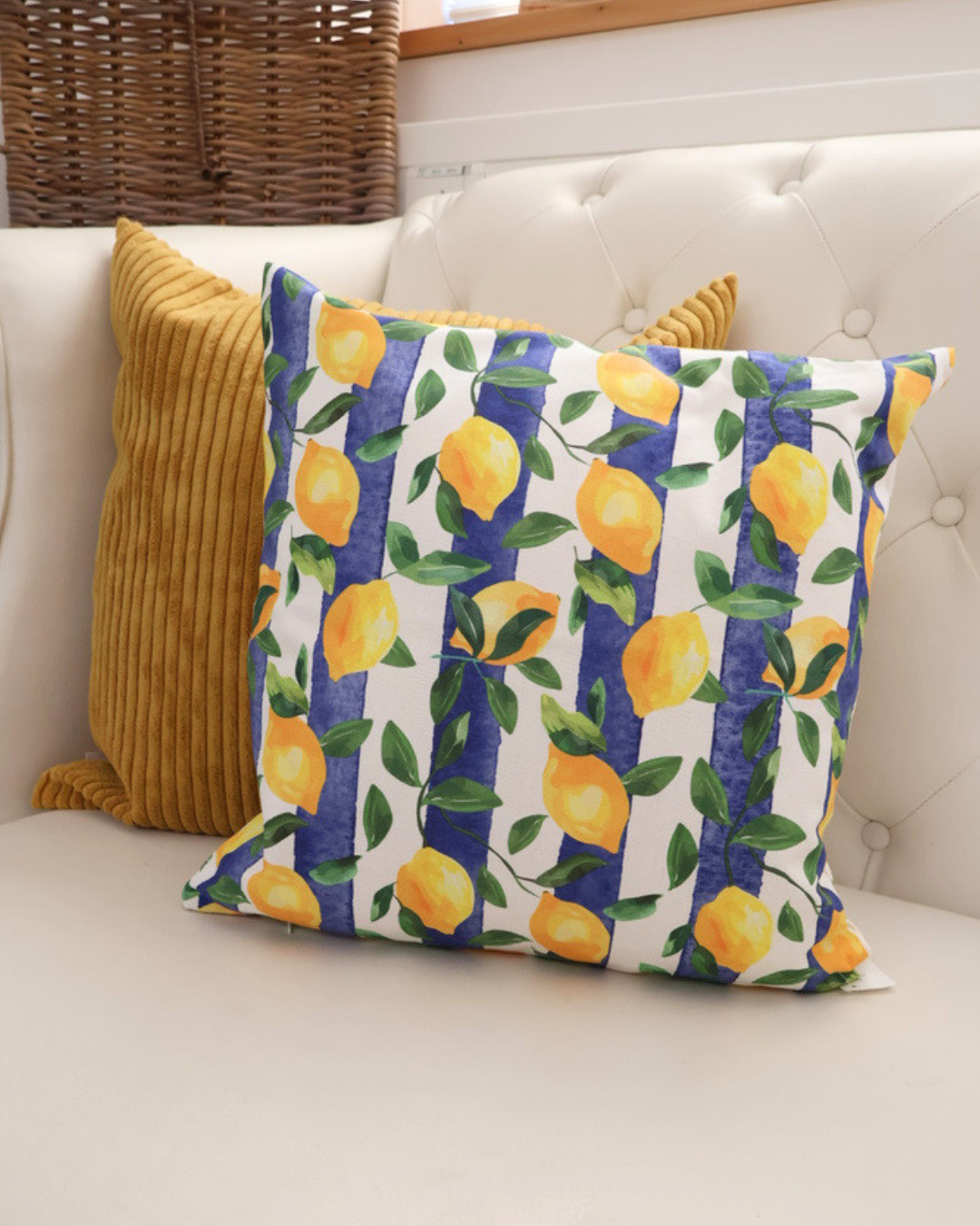 Dekoiertes Bild des Zitronen Kissens "Lemontree". Das Motiv besteht aus Blättern und Zitronen vor einem Blau Weiß gestreiften Hintergrund. Das Kissen liegt neben dem "King" Kissen auf einem weißen Sofa.