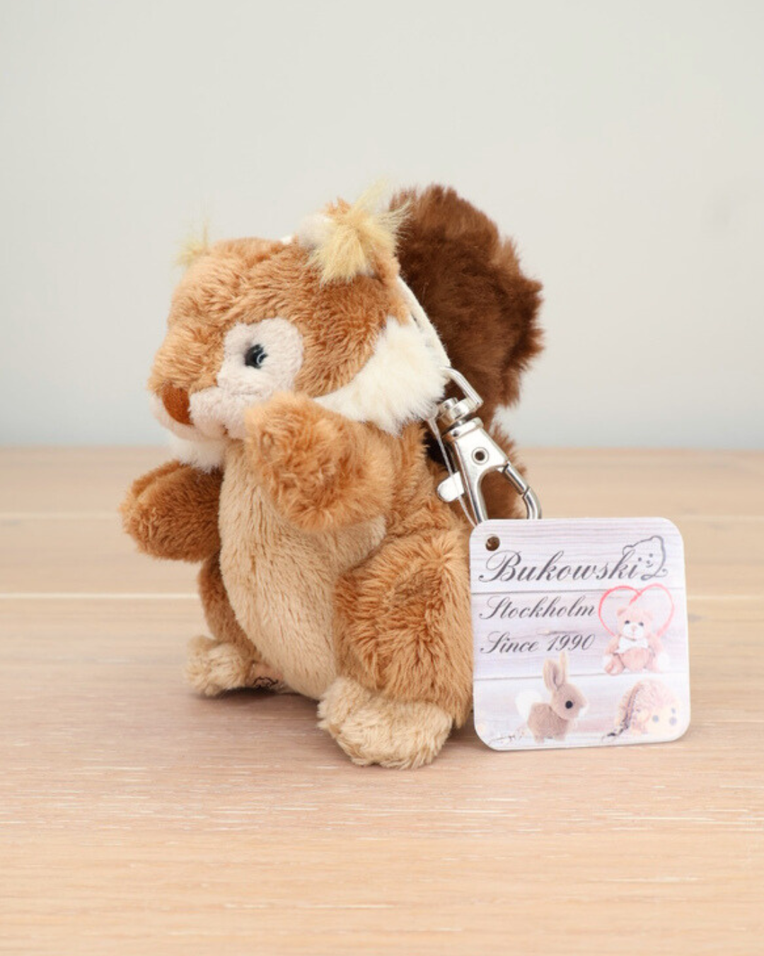 Ein braunes Eichhörnchen von Bukowski sitzt auf einem Holztisch. Der Metallkarabiner und das Markenetikett sind deutlich sichtbar.