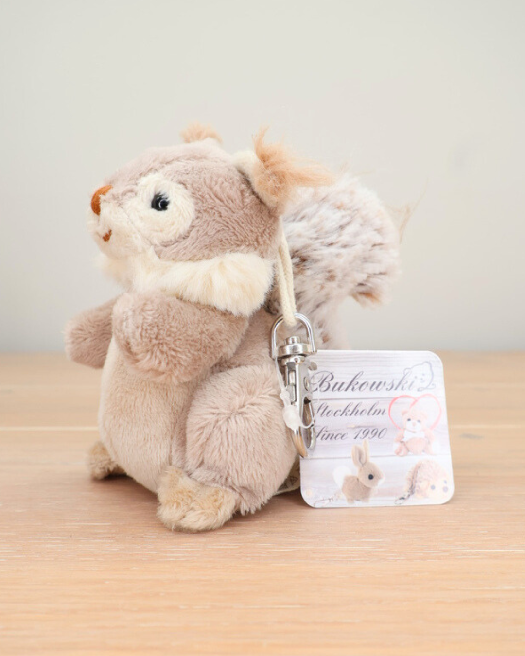 Ein beiger Eichhörnchen-Schlüsselanhänger von Bukowski sitzt auf einem Holztisch. Der Karabiner und das Etikett „Bukowski Stockholm Since 1990“ sind zu sehen.