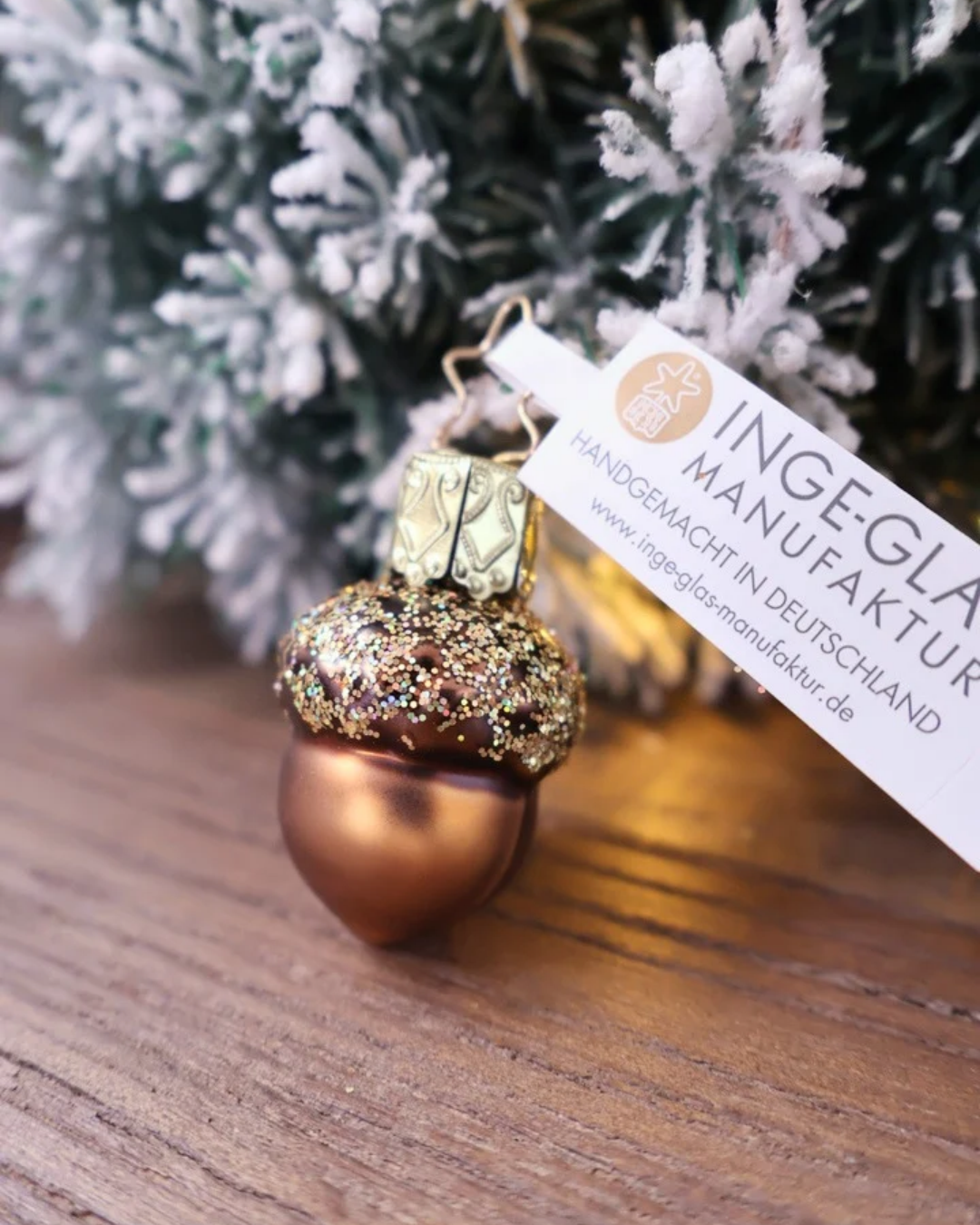 Schräge Ansicht des kleinen weihnachtlichen Eichel Ornaments von Inge Glas, welches vor weiß gepudertem Tannengrün auf einem Holztisch dekoriert ist.