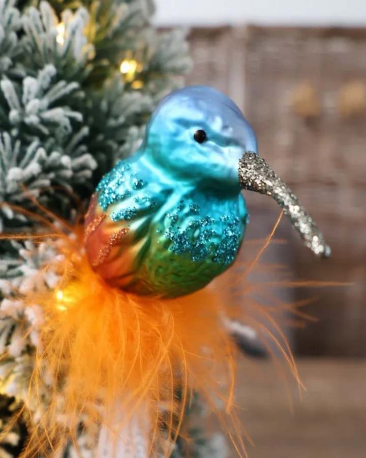 Schrägansicht des exotischen Kolibri Ornaments zum Klemmen von Inge Glas vor dekoriertem Tannengrün. Der aus Glas gefertigte Kolibri sitzt auf echten orangefarbenen Federn. Der Vogel selbst hat orange schimmernde Flügel mit orangefarbenen Glitzer Highlights und blaue Brust- und Kopffedern mit blauen Glitzer Highlights.