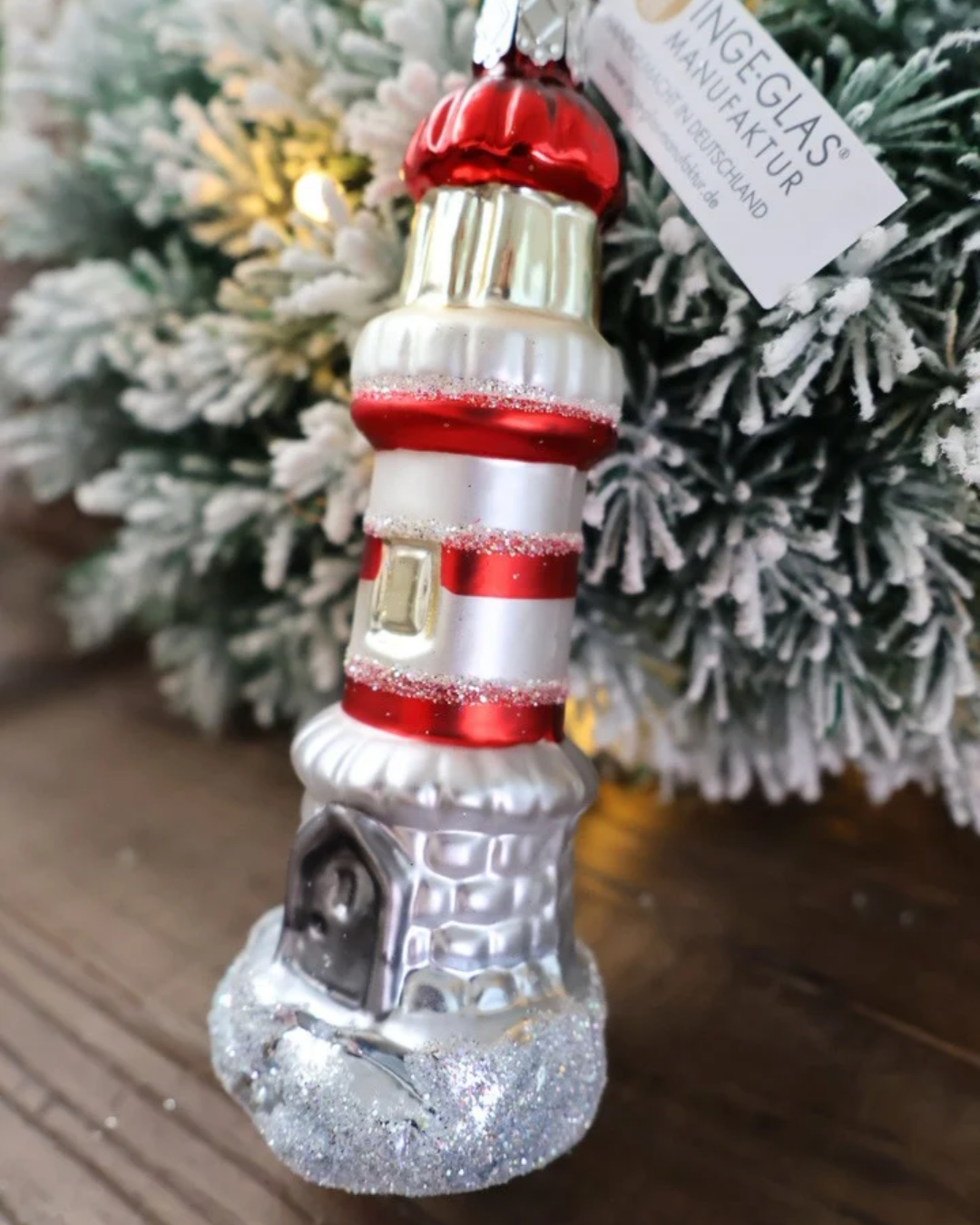 Schräge Ansicht des weihnachtlichen Leuchtturm Ornaments von Inge Glas, welches vor weiß gepudertem Tannengrün auf einem Holztisch dekoriert ist.